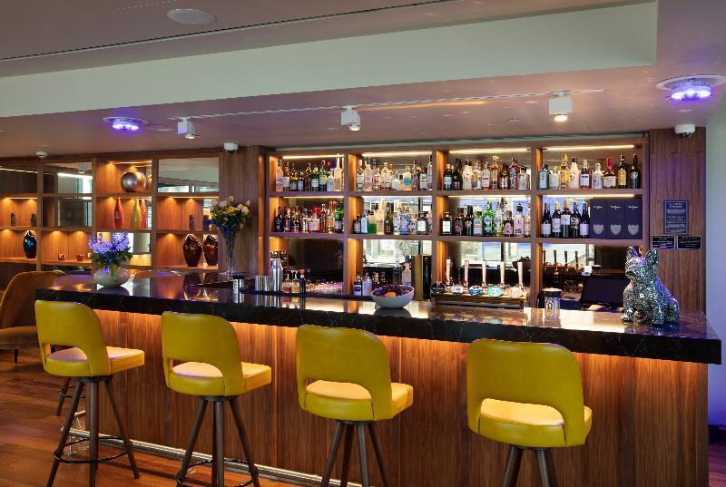 Tower Suites By Blue Orchid Лондон Екстер'єр фото The photo shows a modern bar area. In the foreground, there are several yellow bar stools lined up in front of a sleek black countertop. Behind the bar, there is a well-stocked shelf displaying a variety of bottles, including spirits and liqueurs, il