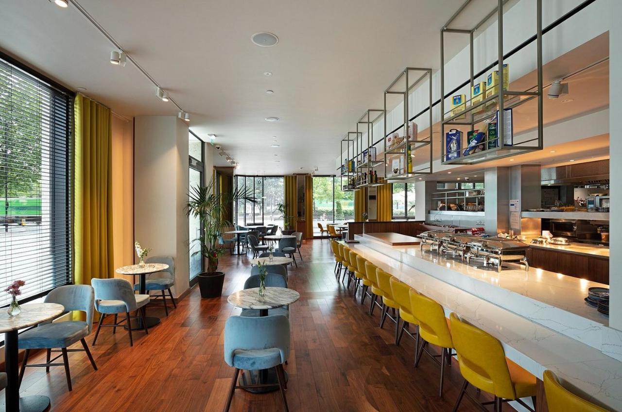 Tower Suites By Blue Orchid Лондон Екстер'єр фото The photo shows a modern restaurant interior. It features a spacious layout with large windows allowing natural light to flood the space. The decor includes a combination of wooden flooring and colorful furniture—blue and yellow chairs arranged aroun