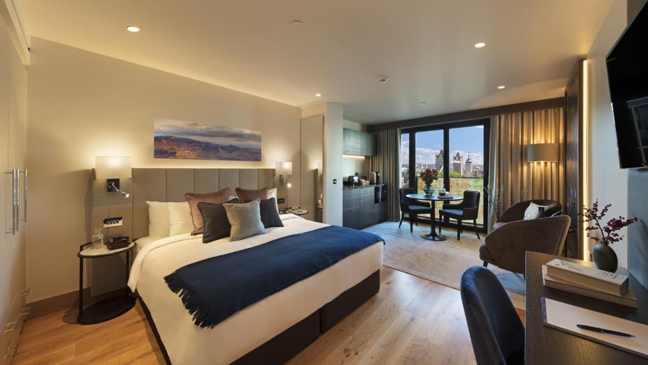 Tower Suites By Blue Orchid Лондон Екстер'єр фото The photo shows a modern hotel room. It features a large bed with a dark blue throw blanket and several decorative pillows. The walls are painted in neutral tones, and there's a landscape painting above the bed. To one side, there is a small kitchene