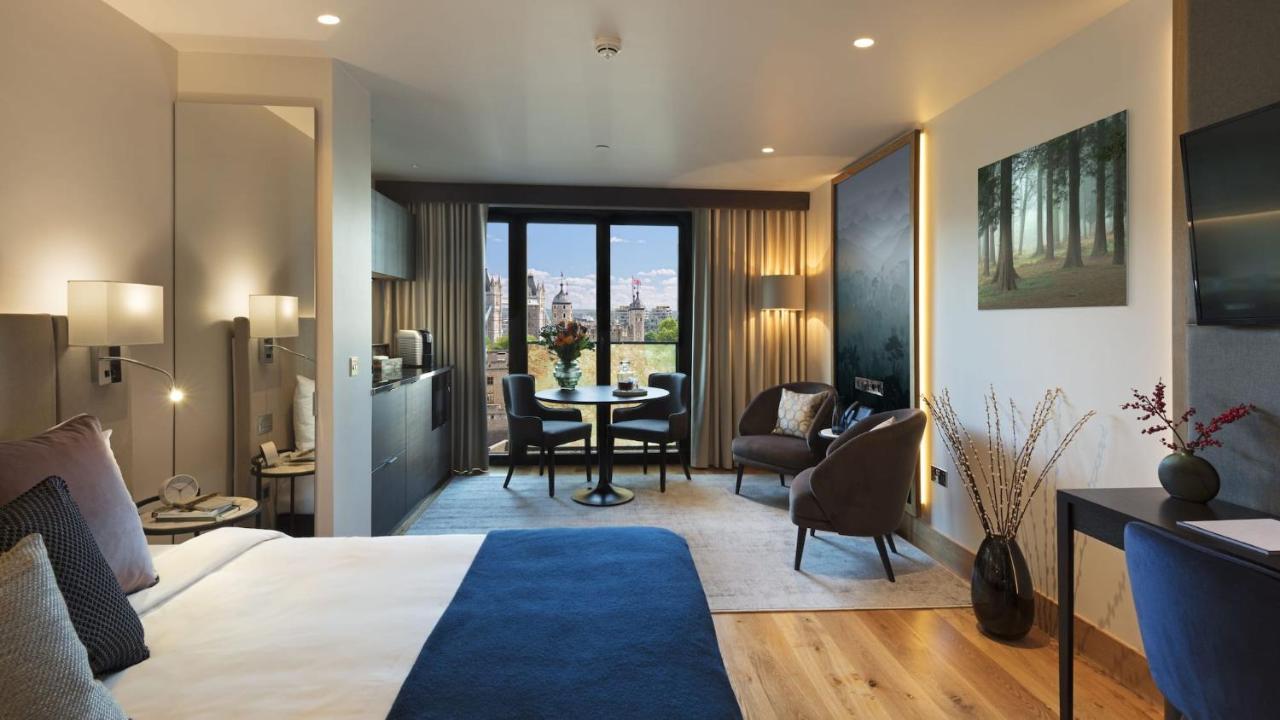 Tower Suites By Blue Orchid Лондон Екстер'єр фото The photo shows a modern hotel room designed with a contemporary aesthetic. On one side, there is a large bed with a blue blanket, while the other side features a sitting area with two chairs and a small round dining table. Large windows let in natur