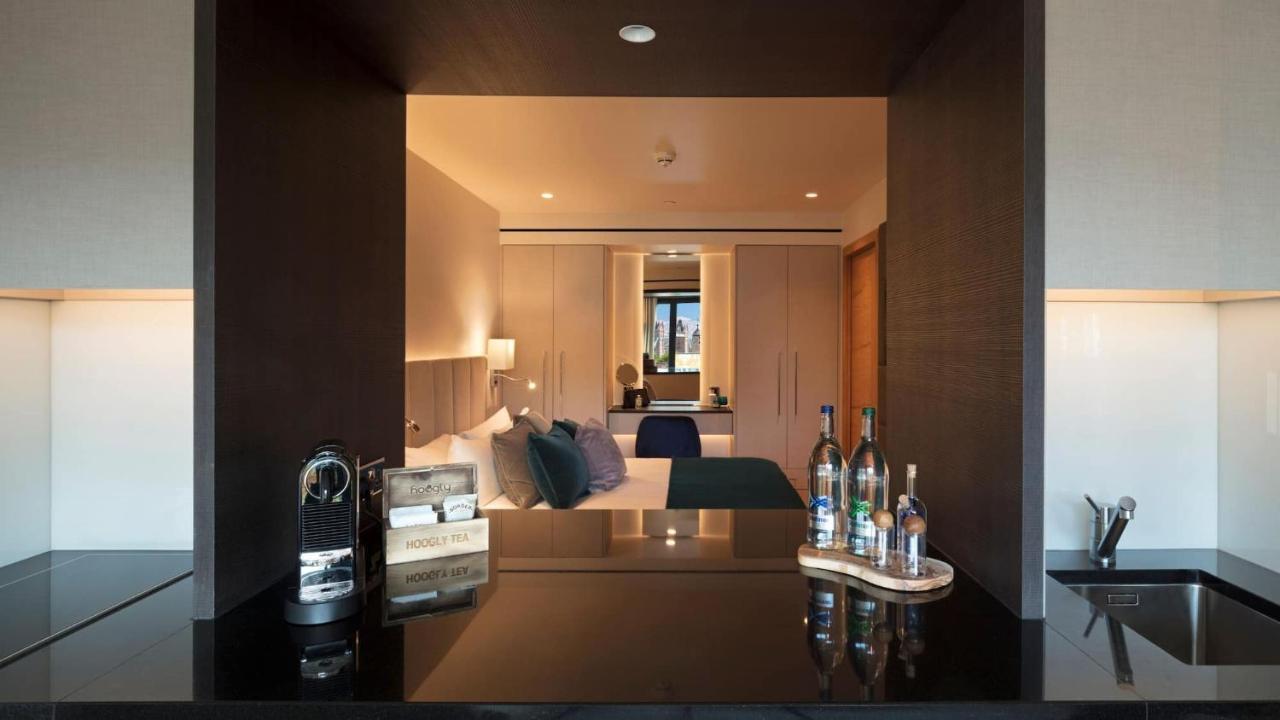 Tower Suites By Blue Orchid Лондон Екстер'єр фото The image shows a modern hotel room with a stylish design. In the foreground, there is a dark countertop with several bottles displayed, likely including spirits and water. Just beyond the countertop, you can see the bedroom area, which has light-col