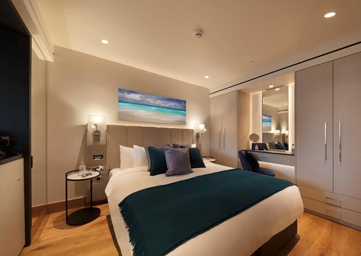 Tower Suites By Blue Orchid Лондон Екстер'єр фото The photo shows a modern hotel room featuring a double bed with a white duvet and a dark teal throw blanket. There are decorative pillows in shades of blue and purple on the bed. To one side, there's a round bedside table with a small lamp. A large m