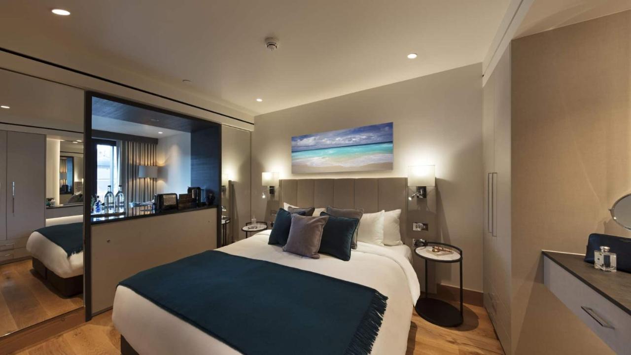 Tower Suites By Blue Orchid Лондон Екстер'єр фото The photo shows a modern hotel bedroom featuring a large bed with a light-colored headboard and a deep blue throw at the foot. The bed is adorned with multiple decorative pillows. On the wall above the bed, there is a colorful painting depicting a be