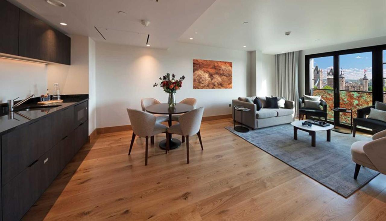 Tower Suites By Blue Orchid Лондон Екстер'єр фото The photo shows a modern apartment interior. On the left side, there is a kitchen area with dark cabinetry, while on the right side, a comfortable living area is visible. In the center, a round dining table is set with four chairs and a vase of flowe