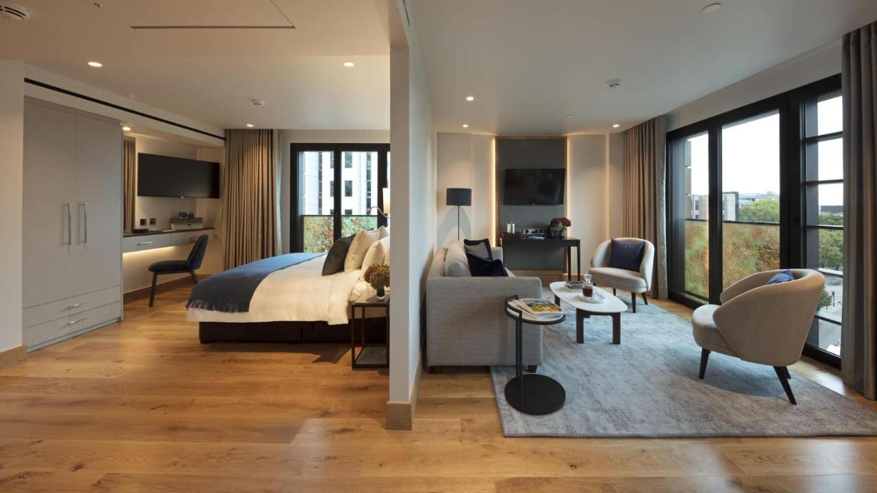 Tower Suites By Blue Orchid Лондон Екстер'єр фото The photo shows a modern hotel room or suite. On one side, there's a bed with a blue and white duvet and pillows, accompanied by a bedside table with a lamp. Opposite the bed, there's a living area featuring a cozy gray sofa, a small round coffee tab