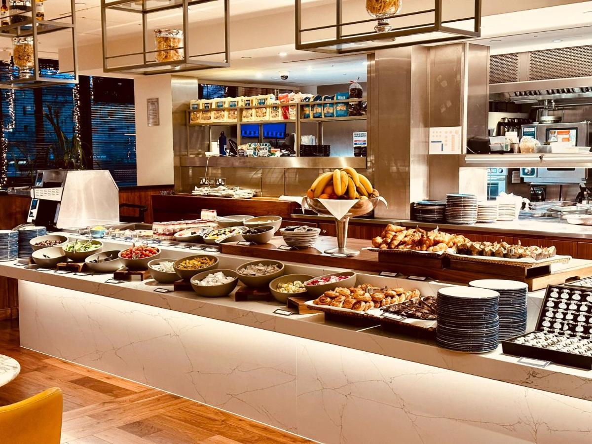 Tower Suites By Blue Orchid Лондон Екстер'єр фото The photo depicts a buffet-style setup, likely in a restaurant or a hotel dining area. In the foreground, there are multiple bowls and platters filled with a variety of food items, including salads, fruits, pastries, and other dishes. There is a long