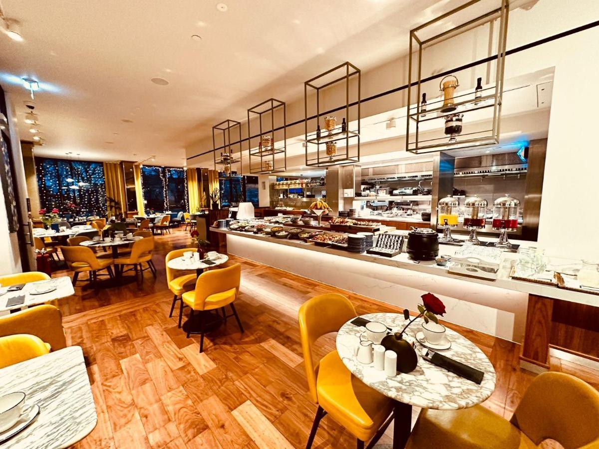 Tower Suites By Blue Orchid Лондон Екстер'єр фото The photo depicts a modern restaurant interior with a spacious layout. There are several round tables, each adorned with marble tabletops and stylish yellow chairs. A buffet area is visible in the background, featuring various food setups and kitchen
