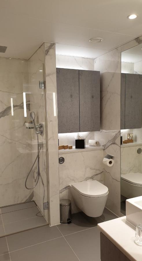 Tower Suites By Blue Orchid Лондон Екстер'єр фото The photo shows a modern bathroom design featuring elegant marble walls. On one side, there is a transparent shower enclosure with a showerhead and controls visible. Opposite the shower, there is a toilet, and above it are some sleek cabinets. The sp