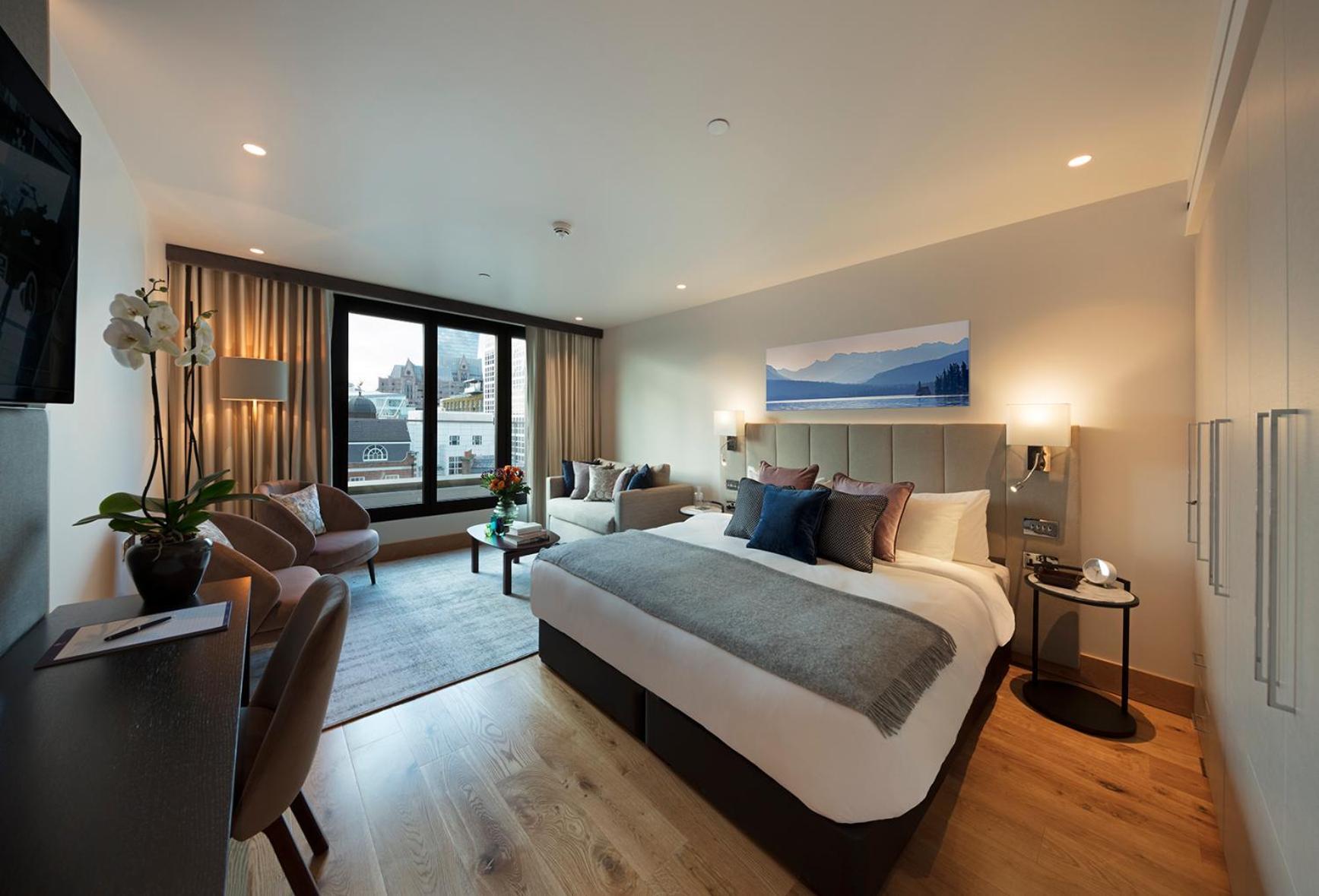 Tower Suites By Blue Orchid Лондон Екстер'єр фото The photo shows a modern hotel room. In the center, there is a large bed with stylish bedding and pillows in various shades. To one side, there are two comfortable chairs and a small table, creating a cozy seating area. A large window offers a view o